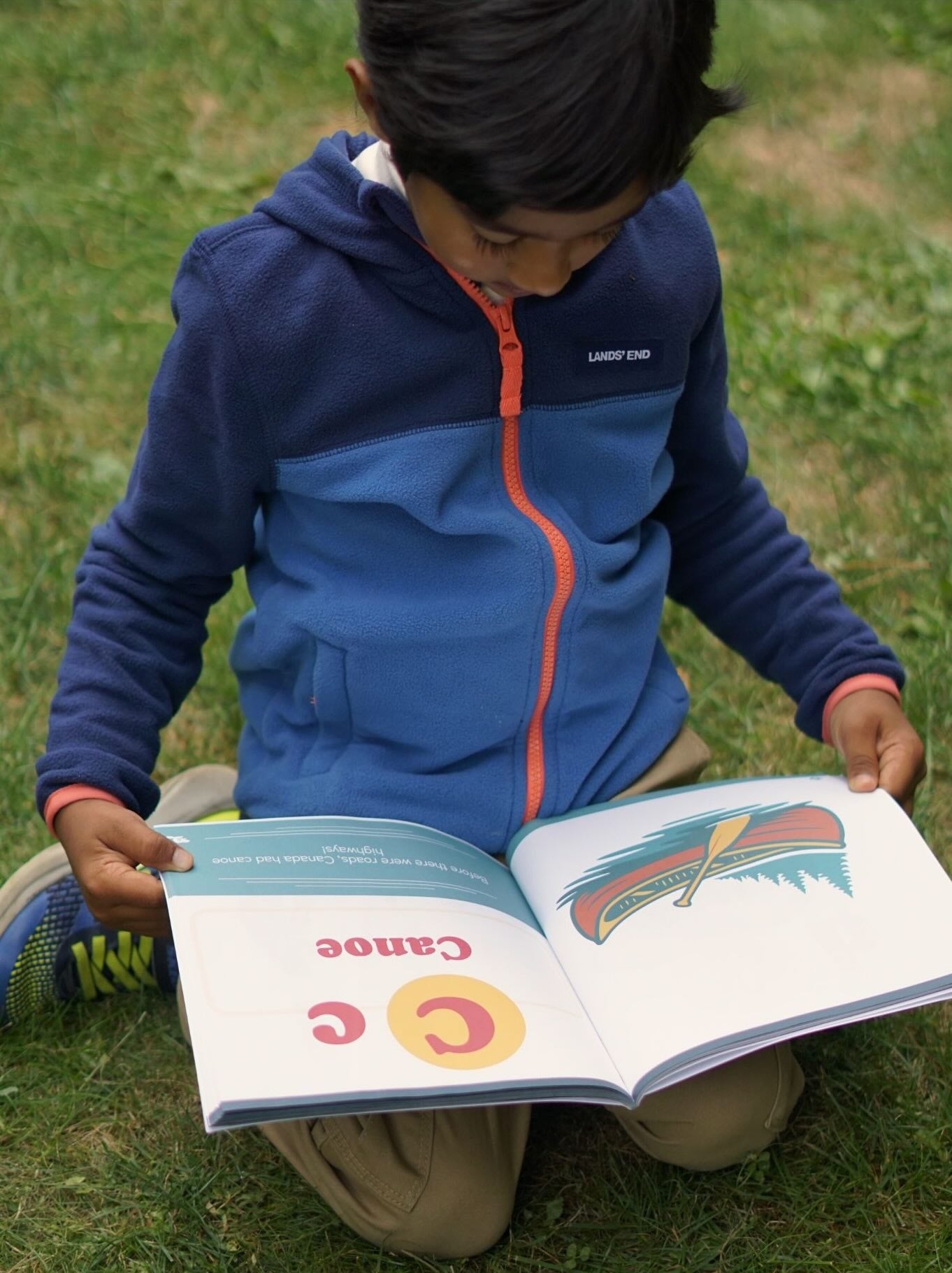 Child reading the book From Aurora to Zamboni, An ABC Journey Through Canada, outdoors on grass