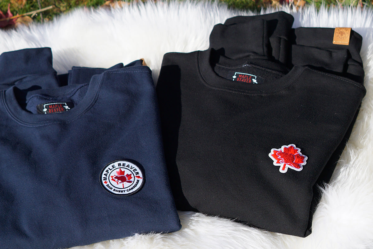 Two folded Maple Beaver Canadian made sweatshirts, one navy and one black, with embriodery on a fluffy white surface.